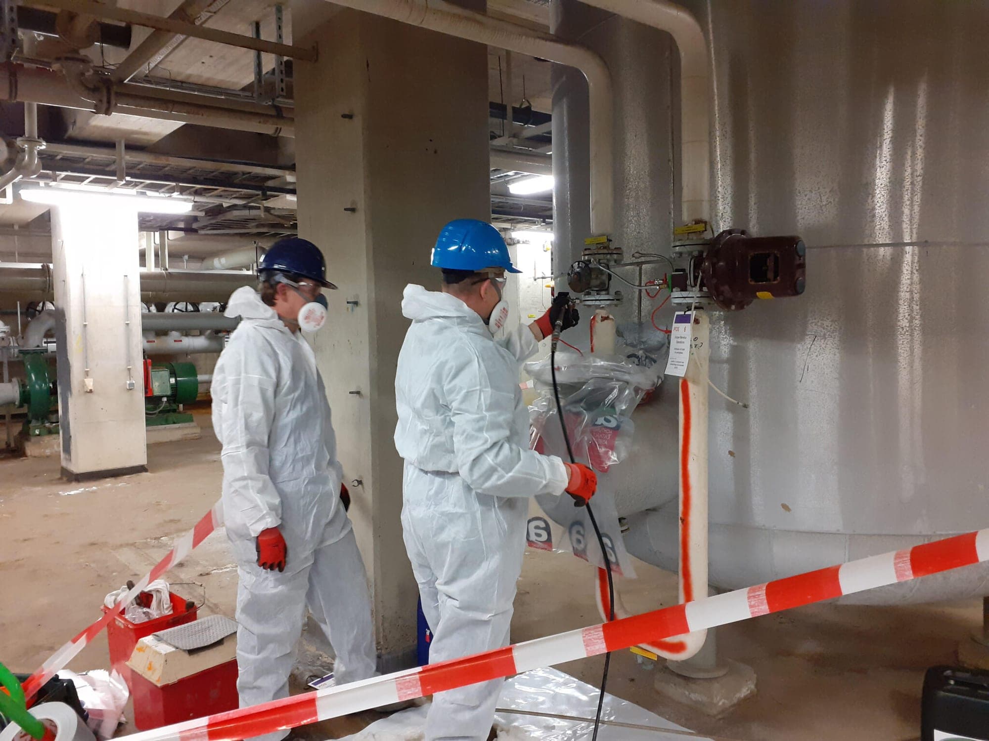 Twee saneerders in beschermende pakken en met adembescherming werken achter afzetlint aan een industriële installatie
